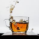Dynamic capture of tea splashing in a transparent glass mug, creating a dramatic visual effect against a white backdrop.